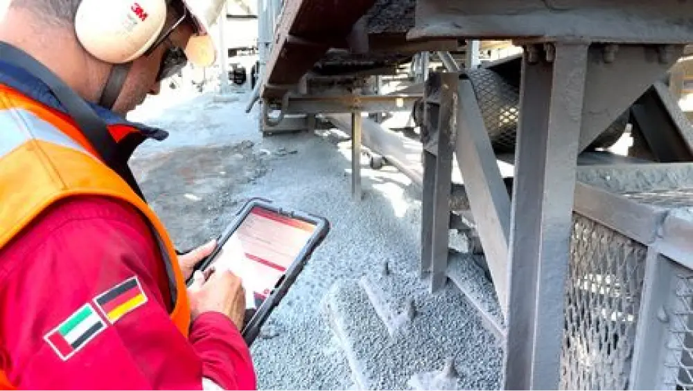 Mitarbeiter in roter Schutzkleidung prüft eine Anlage mit Tablet am Förderband.